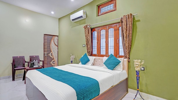 Un dormitorio con cama, dos sillas, una ventana con cortinas, un aire acondicionado y un florero con flores.