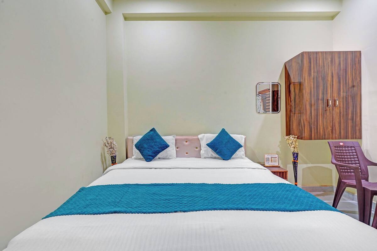 A hotel room with a bed, a wooden wardrobe, a desk with a chair, and a small table with a vase.