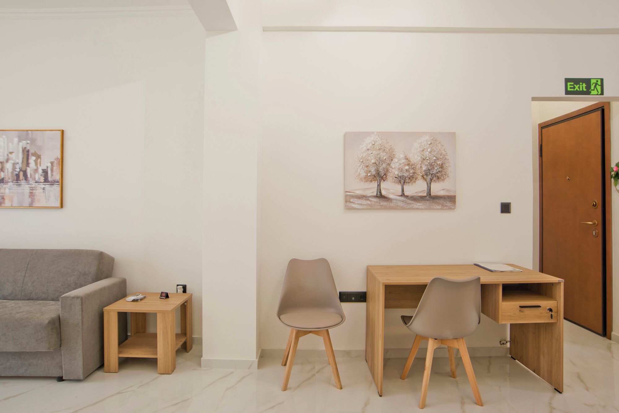 A room with a grey sofa, a wooden desk with two chairs, and a painting of trees.
