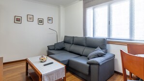 Un salon avec un canapé gris, une petite table sur laquelle se trouve une télécommande et une fenêtre avec des rideaux.