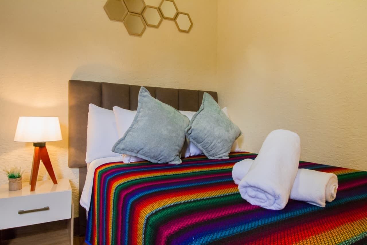A neatly made bed with a colorful blanket, two pillows, and rolled towels on a white nightstand with a lamp and a small plant.