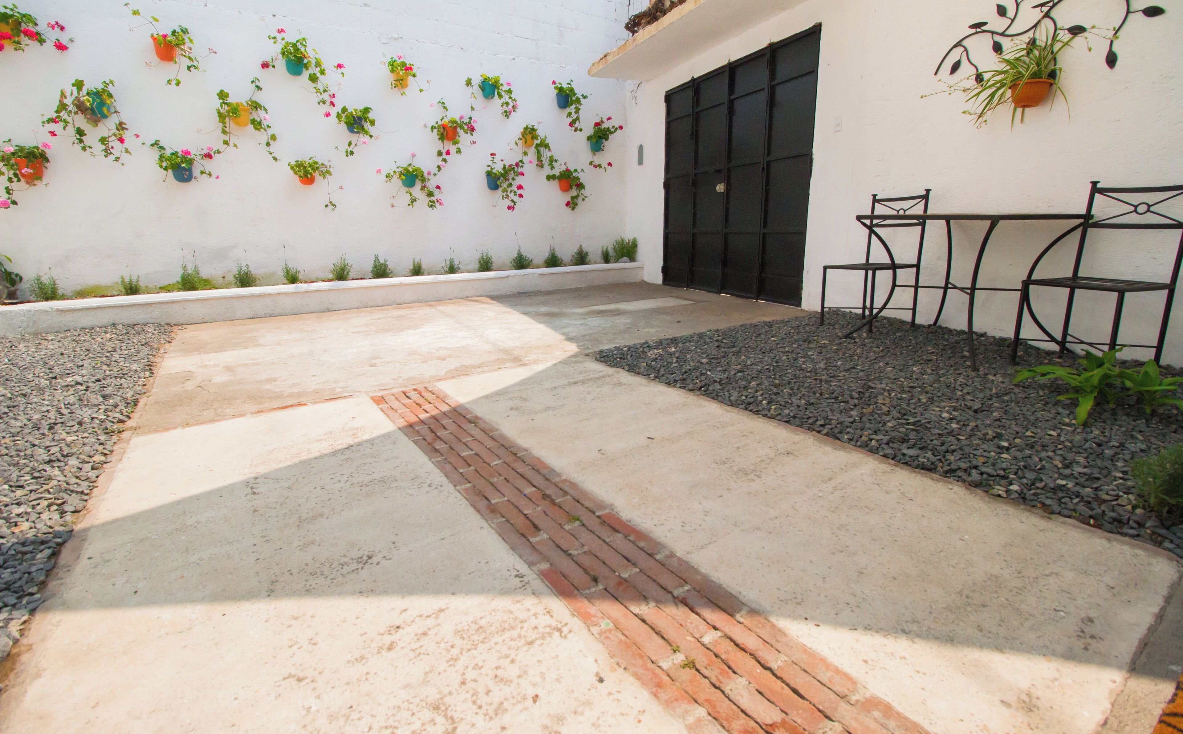 A patio with a concrete floor, a brick pathway, a black door, and a white wall with hanging plants.
