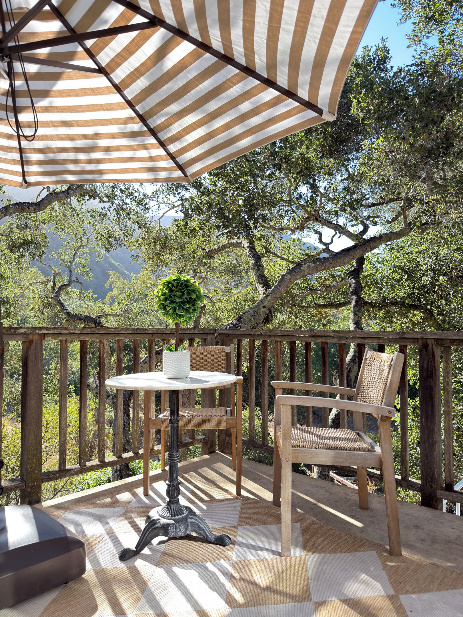 Un patio exterior con una mesa y sillas, un paraguas a rayas y vistas a árboles y montañas.