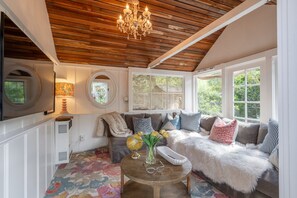 A cozy room with a wooden ceiling, a round table with a vase of flowers, a sofa with various cushions, and a large window providing natural light. - Private River Access & Walk to Village! (Carmel Valley)