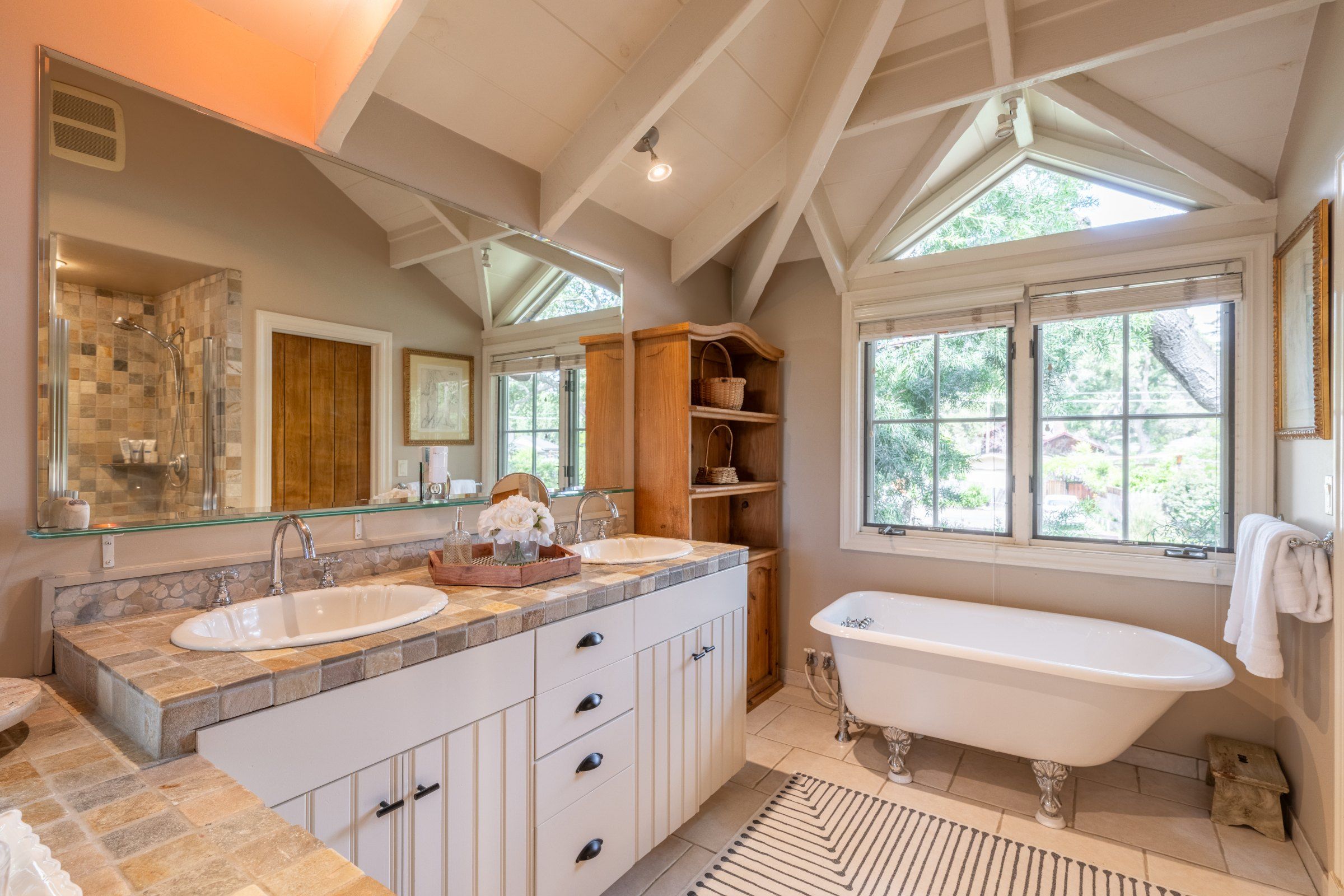 A bathroom with a freestanding bathtub, two sinks, a large mirror, and a window with a view of greenery.