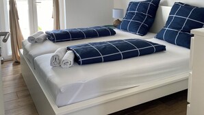 A double bed with white bedding and blue pillows, a bedside table with a lamp, and a window with beige curtains.