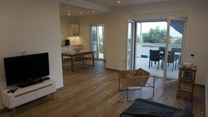 A modern living room with a flat-screen TV, a sofa, a dining area with a table and chairs, and a balcony with outdoor seating.