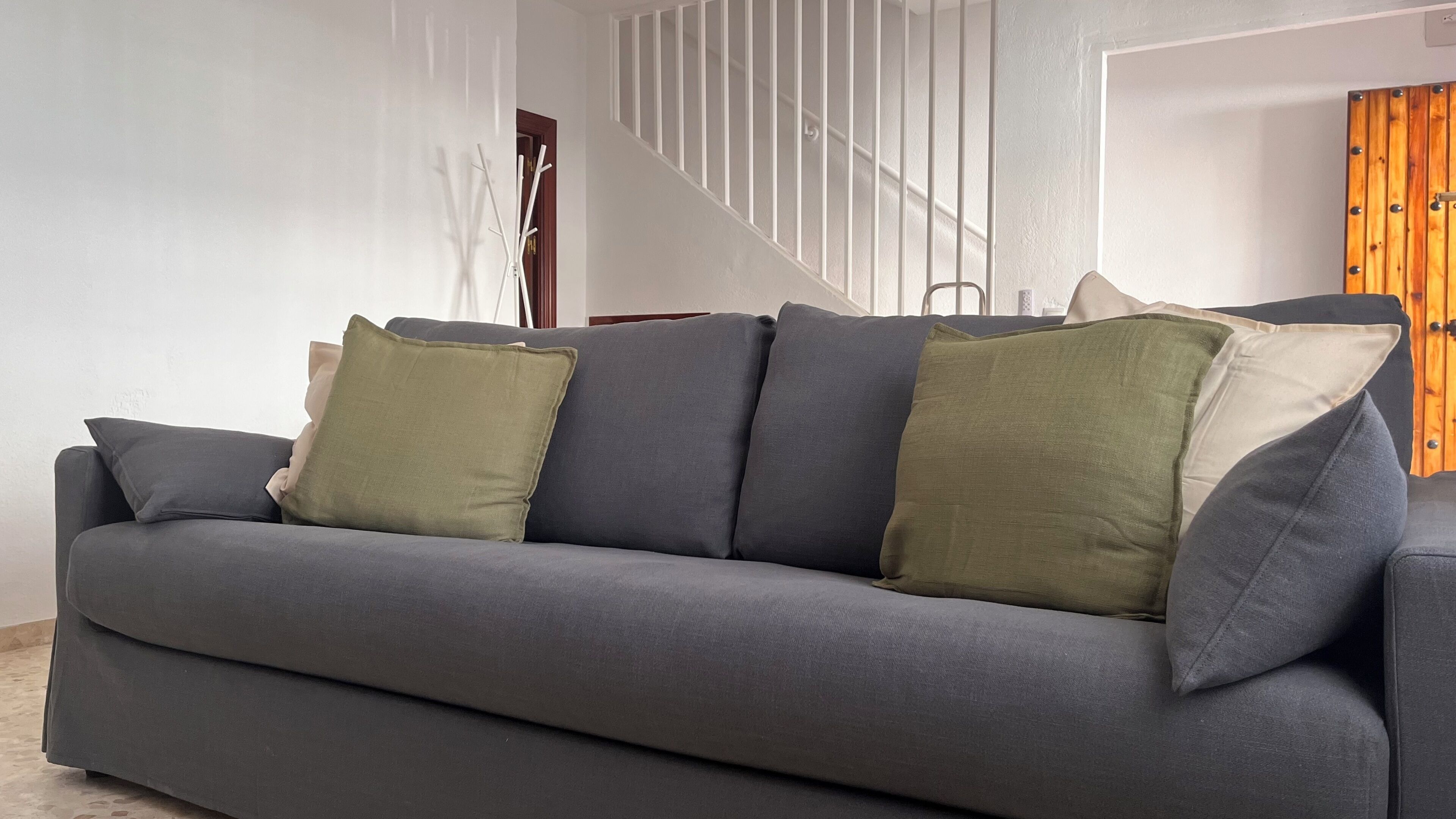 Un canapé gris agrémenté de plusieurs coussins, dans une pièce dotée d’un escalier et d’une porte en bois.