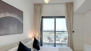 A hotel room with a bed, a chandelier, a large window with curtains, and a view of the outside. - Vogue | Olivz Residence by Danube Near Dragon Mart (Dubai)