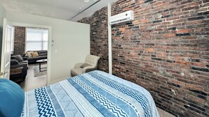 Une pièce avec un mur en briques, un ventilateur de plafond, un lit avec une literie à motifs, une chaise et une fenêtre avec des stores.