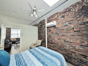 A room with a brick wall, a ceiling fan, a bed with patterned bedding, a chair, and a window with blinds. - Popular Queen Suite w/ Parking – SLU 201 (St. Louis)