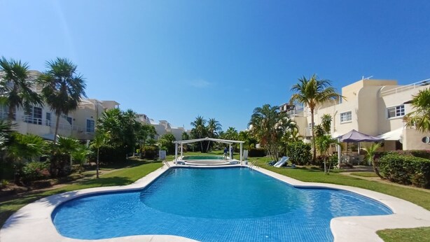 Une piscine dotée d’une terrasse blanche, entourée de palmiers et surplombée d’un ciel bleu azur.