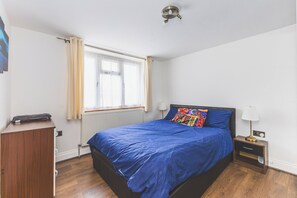 A bedroom with a bed, a nightstand, a lamp, a window with curtains, and a radiator. - Bright 2BDR Apt in Southall with Free Parking (Greater London)