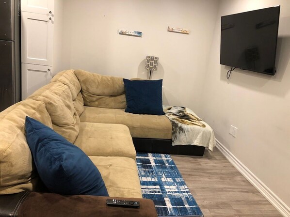 Un salon avec un canapé d’angle beige, un coussin bleu, une télécommande posée sur un pouf marron, un tapis à motifs et un téléviseur à écran plat fixé au mur.