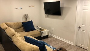 Una sala de estar con un sofá seccional, un televisor de pantalla plana montado en la pared, una puerta y una mesita con un control remoto.