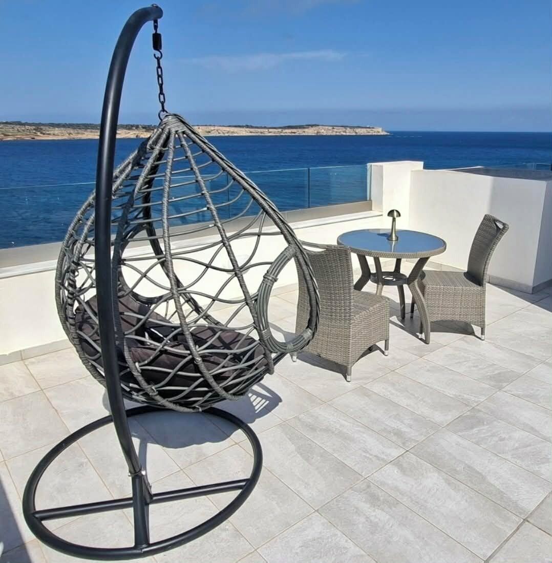En hängstol, ett litet runt bord och två stolar på en terrass med utsikt över havet.