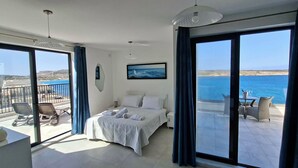 Una camera da letto con un letto, una scrivania e una sedia, un balcone con vista sul mare e un'area pranzo con tavolo e sedie.
