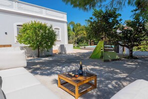 Un edificio blanco con un patio, una mesa de madera con una botella y vasos, y una carpa verde.