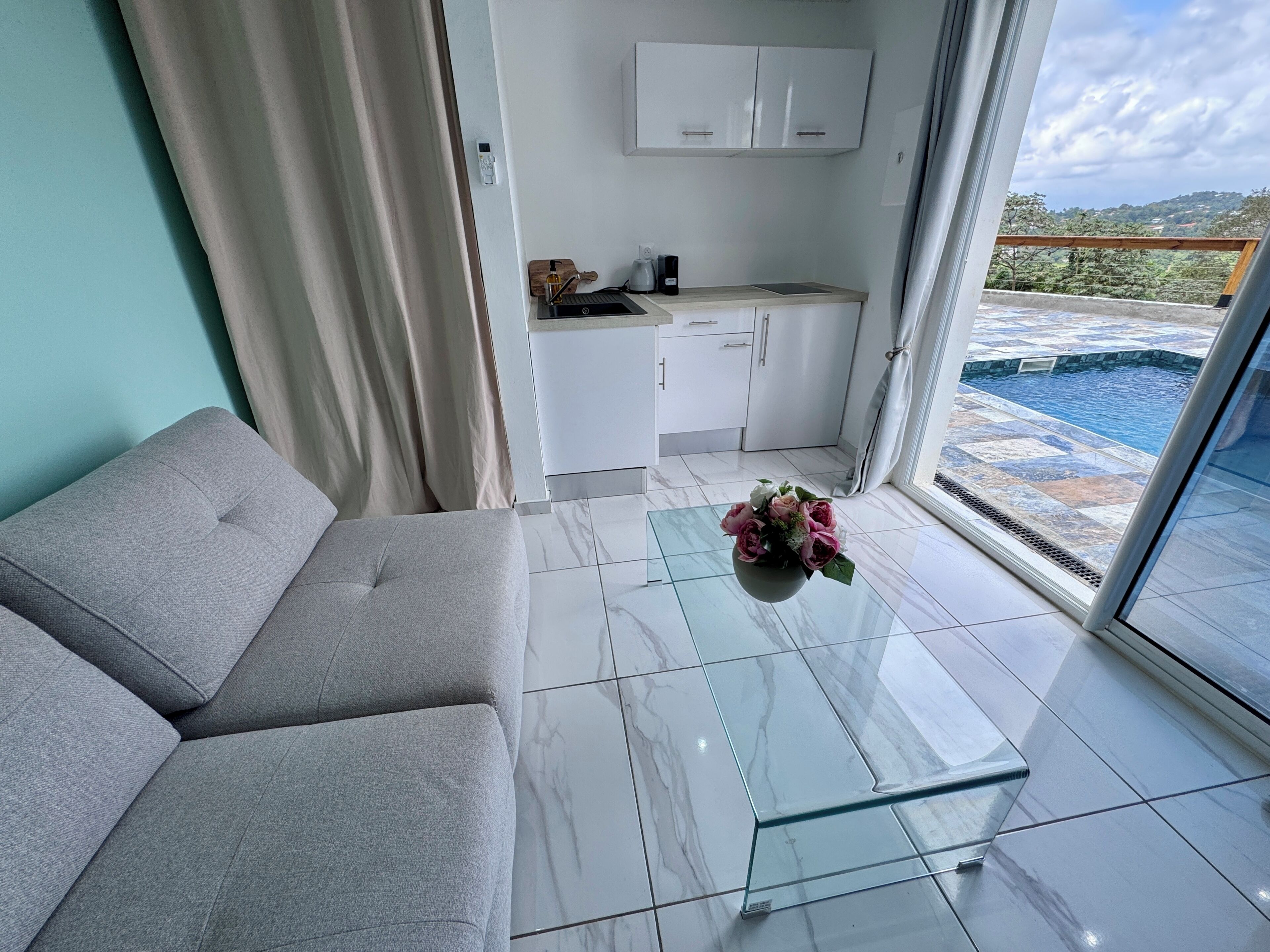 A modern living room with a sofa, a glass coffee table with flowers, a kitchen area, and a view of a swimming pool.