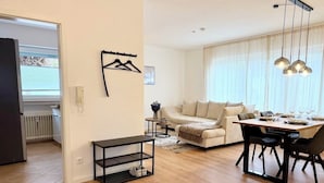 A living room with a sofa, a dining table with chairs, a coffee table, a hanging light fixture, and a coat rack. - 3BR | parking, WLAN, home office, balcony, view, fully equipped. Kitchen (Bad Mergentheim)