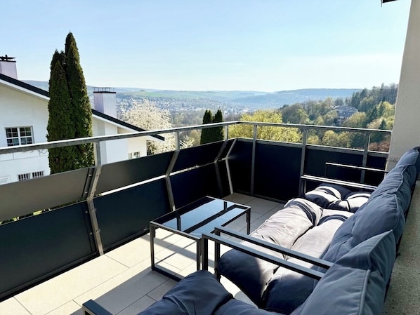 Eine Dachterrasse mit einer Sitzecke, einem Glastisch und Blick auf die Stadtlandschaft.