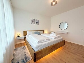 Ein ordentlich bezogenes Bett mit weißen Leinentüchern, ein Nachttisch mit Lampe, ein runder Spiegel an der Wand und eine kleine Ablage mit gerollten Handtüchern.
