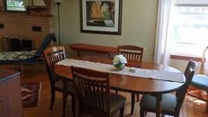 A dining area with a wooden table, chairs, a vase with flowers, a framed painting, and a window with curtains.