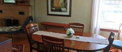 A dining area with a wooden table, chairs, a vase with flowers, a framed painting, and a window with curtains.