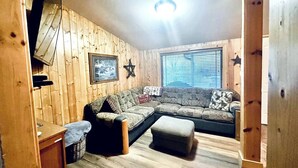 A cozy living room with a sectional sofa, a small ottoman, and a wooden floor. - Serene 2-bedroom cabin on The Provo River (Provo)