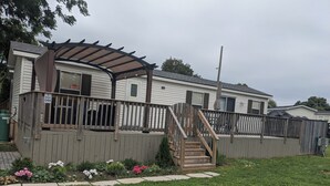 A single-story mobile home with a covered patio, a wooden deck, and a small garden with flowers. - Spacious Corner Lot Cottage @ Sherkston Shores (Port Colborne)