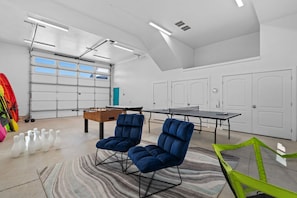 Un grand espace intérieur comprenant une table de ping-pong, deux chaises bleues, une table de baby-foot et une porte de garage.
