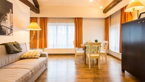Ein Wohnzimmer mit einer Couch, einem Esstisch mit Stühlen, einer dunklen Holzkommode und einem Fenster mit Vorhängen.