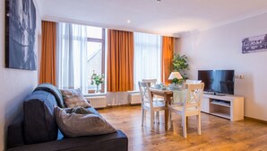 Una sala de estar con sofá, una mesa de comedor con sillas, televisor sobre un mueble bajo y ventanas amplias con cortinas.