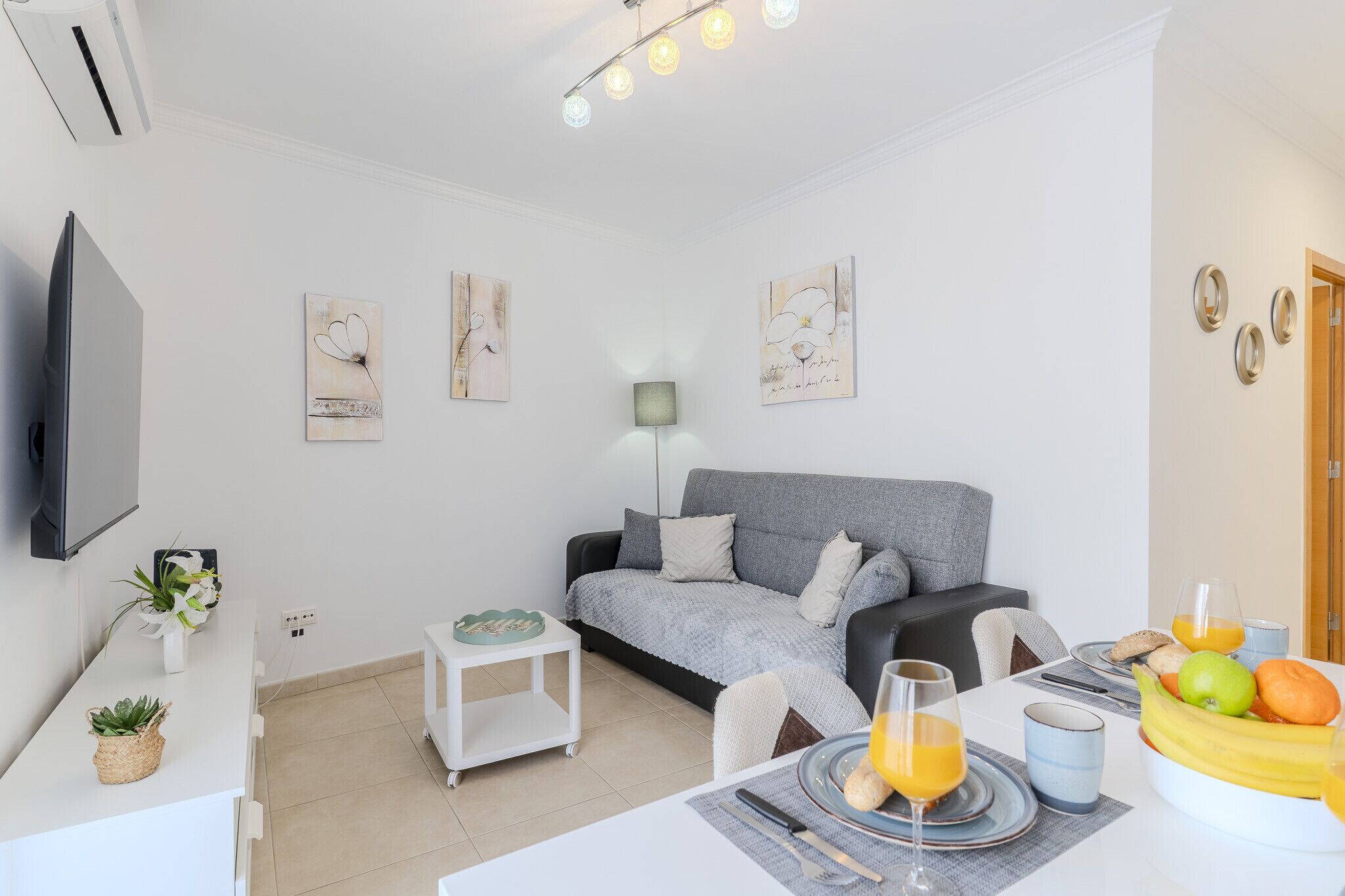 A modern living room with a grey sofa, a dining table with fruit and drinks, a flat-screen TV, and artwork on the walls.