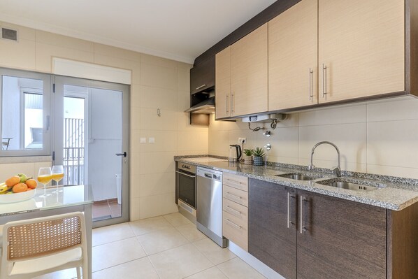 A modern kitchen with a dishwasher, double sink, and a dining area with a table and chairs. - #368 T2 Apartment | Ocean View & Terrace in Albufeira (Albufeira)