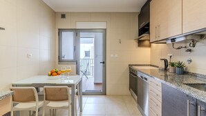 Een moderne keuken met een eethoek, met een glazen tafel, witte stoelen en uitzicht door een open deur naar een balkon.