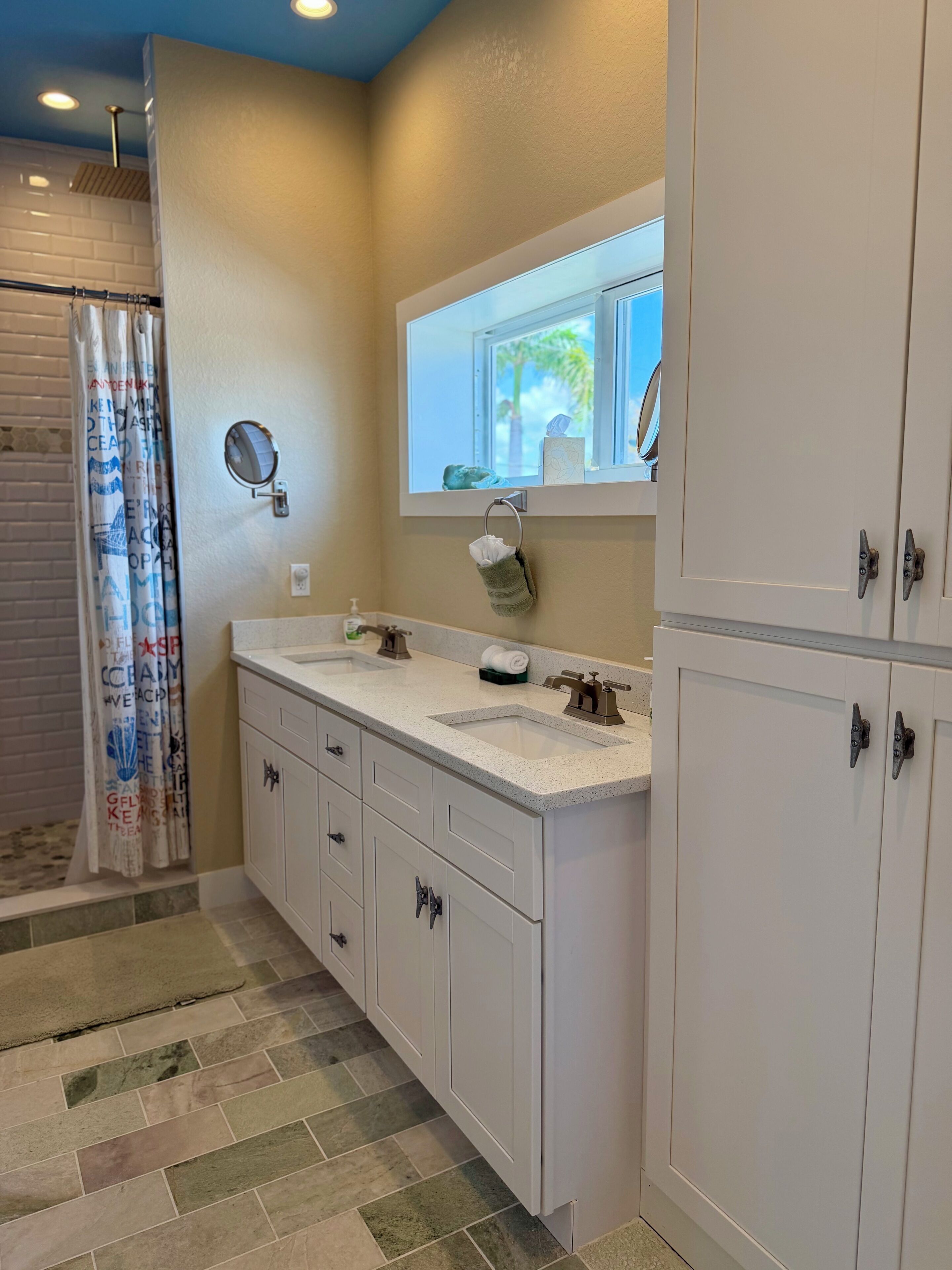 A bathroom with a shower, a sink with a mirror, and a window with a view of palm trees.