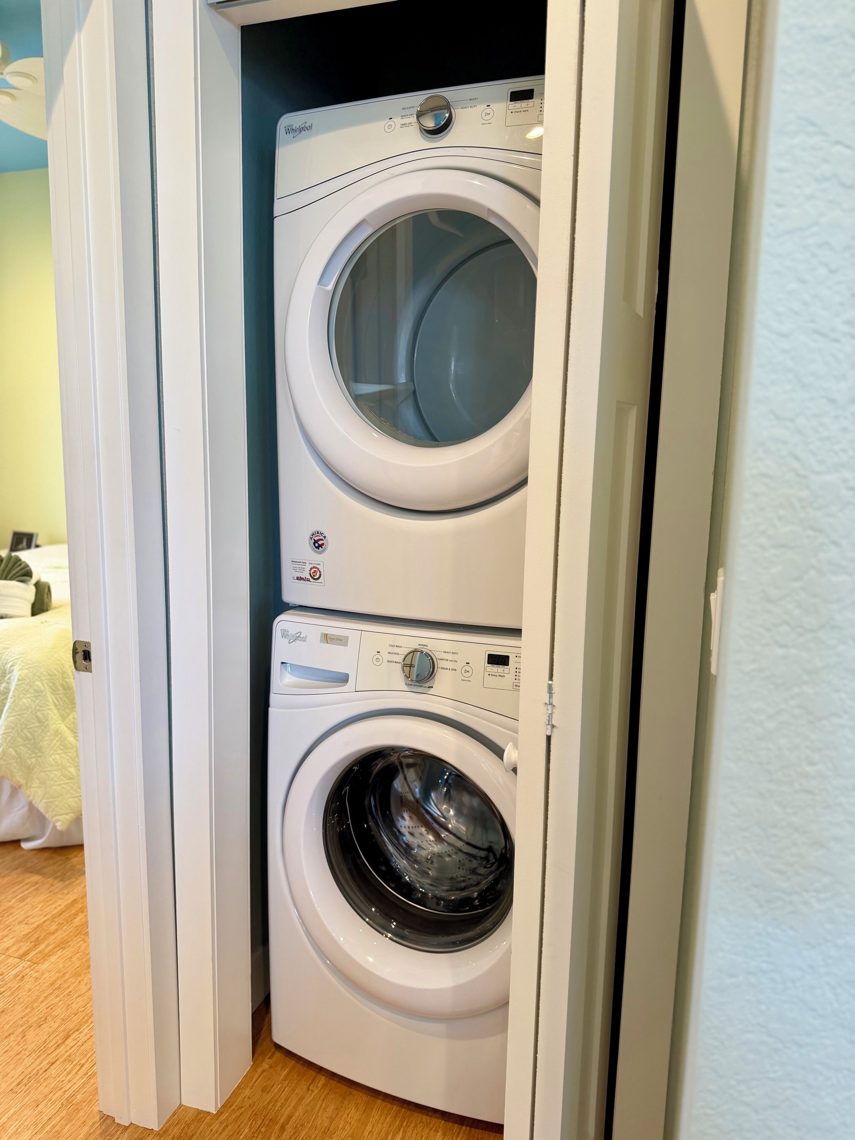 A front-loading washer and dryer unit in a laundry room with a wooden floor and a bed in the adjacent room.