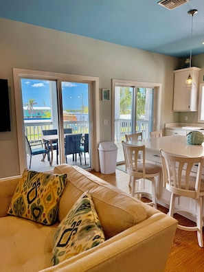 A living room with a sofa, a dining area with chairs, a kitchen with cabinets, and a sliding glass door leading to an outdoor deck. - The Island Shanty (Matlacha)