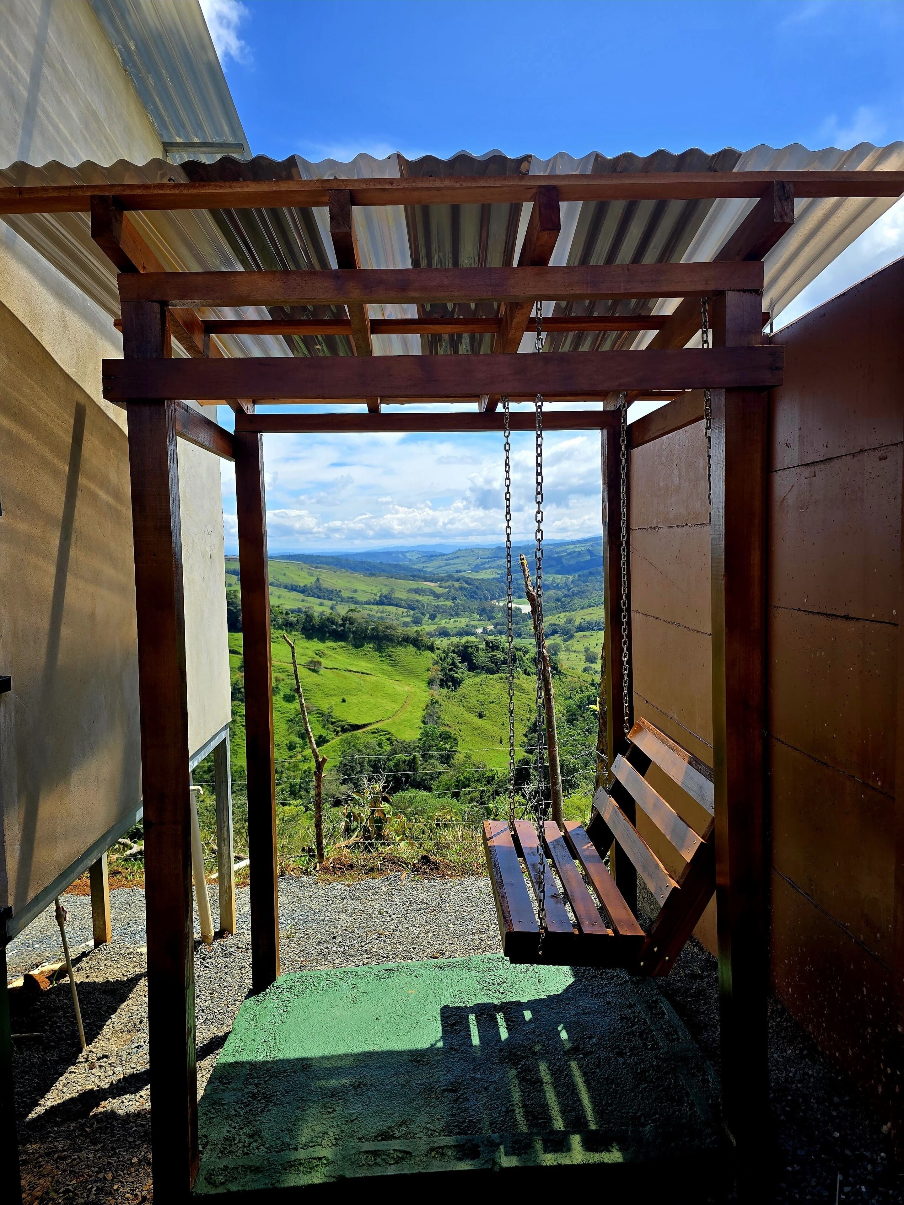 A wooden swing under a corrugated metal roof with a view of green hills.