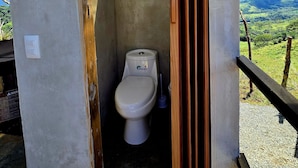 A small, enclosed bathroom with a toilet, a visible toilet brush, and a view of a green landscape through an open doorway. - Dulces Momentos PZ (San Pedro)