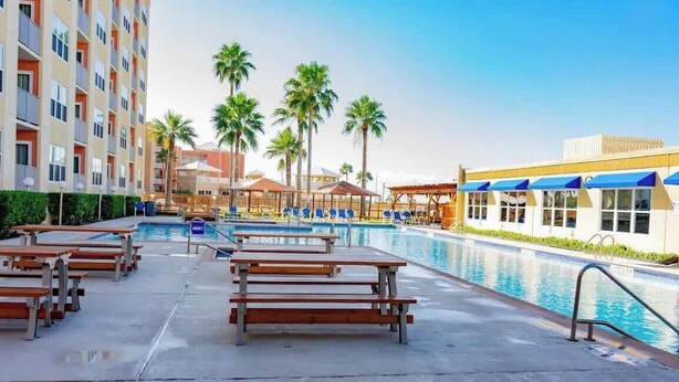 A hotel pool area with multiple lounge chairs and benches, surrounded by palm trees and a multi-story building. - Beach Access Condo - Pool - Hot Tub With BBQ (South Padre Island)