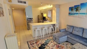 A living room with a grey sofa, a kitchen with a bar area, and a patterned rug. - Beach Access Condo - Pool - Hot Tub With BBQ (South Padre Island)