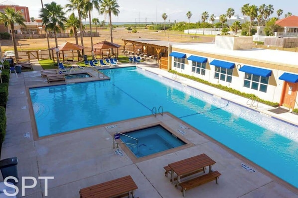 Un área de piscina con sillas de descanso, un jacuzzi y un edificio con toldos azules.