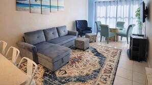 A living room with a grey sofa, patterned ottomans, a dining area with chairs, a TV, and a ceiling fan. - Beach Access Condo - Pool - Hot Tub With BBQ (South Padre Island)