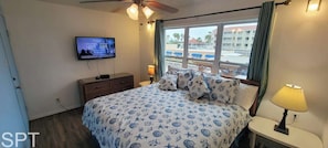 Habitación de hotel con cama, televisión, mesita de noche, ventana con vista a un edificio y palmeras, y ventilador de techo.