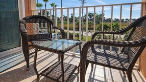 A balcony with wicker chairs and a glass table, overlooking a road with palm trees and a clear sky. - Beach Access Condo - Pool - Hot Tub With BBQ (South Padre Island)