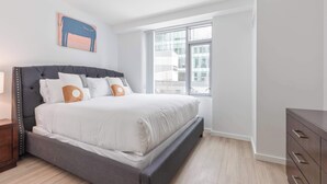 Un dormitorio con cama, mesita de noche, cómoda, ventana y una obra de arte en la pared.
