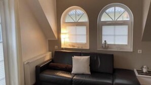 Et værelse med en sort lædersofa, to hvælvede vinduer med skodder, en radiator og et lille bord med en lampe.