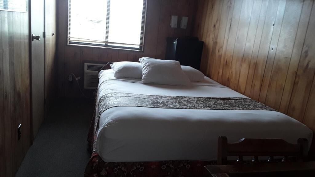 A small, dimly lit room with a single bed, wooden walls, and a window with blinds.